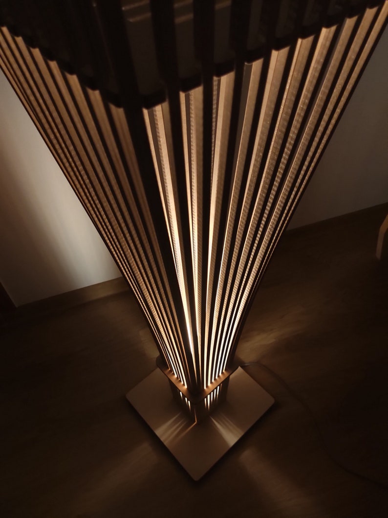 May include: A modern, geometric floor lamp with a square base and a fan-like shade. The lamp is made of light-colored wood and has a warm, inviting glow. The design features vertical wooden slats that create a unique lighting effect.