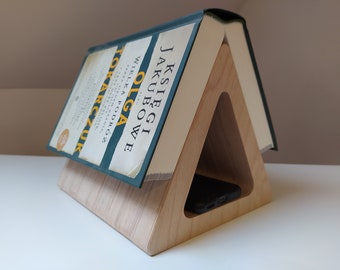 May include: A wooden phone stand with a triangular design. A book titled "OLGA JAKUBOWIE WIELKA PODRÓŻ" by TOKARCZUK is propped on the stand.