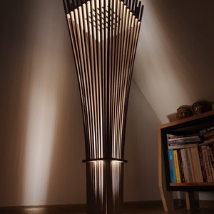 May include: A modern floor lamp with a geometric design. The lamp features vertical wooden slats that create a cage-like structure, with a light source illuminating from within. The lamp is dark brown and stands on a square base.