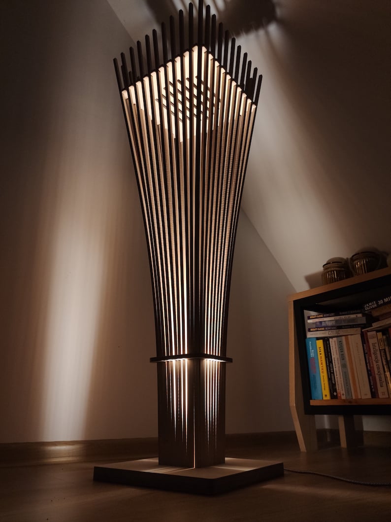 May include: A modern floor lamp with a geometric design. The lamp features vertical wooden slats that create a cage-like structure, with a light source illuminating from within. The lamp is dark brown and stands on a square base.