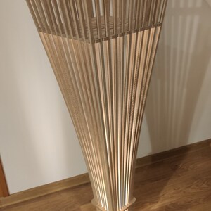 May include: A wooden floor lamp with a unique design. The lamp features a square base and a rectangular midsection, with a flared top made of vertical wooden slats. The lamp is lit from within, creating a warm glow.