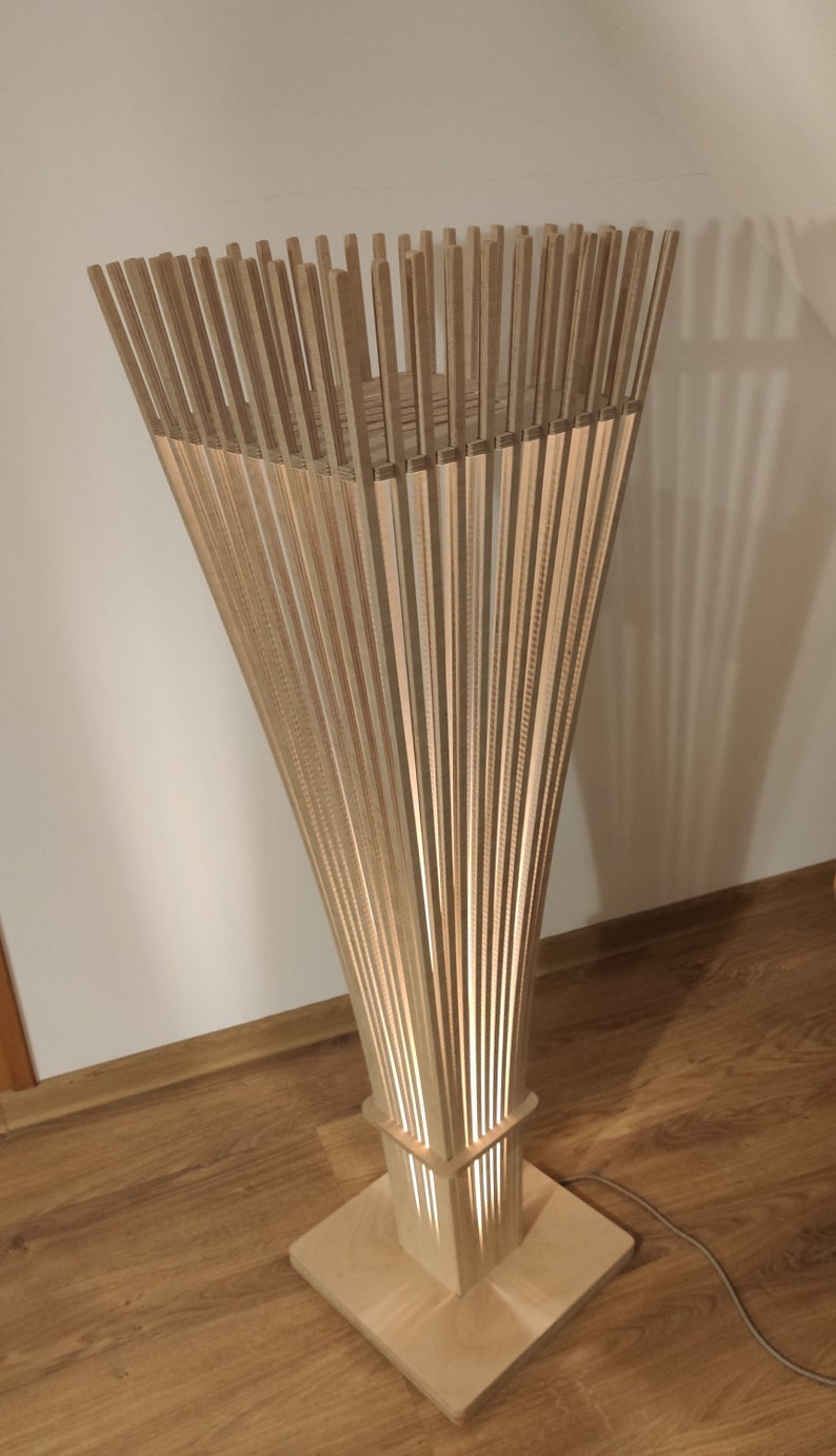 May include: A wooden floor lamp with a unique design. The lamp features a square base and a rectangular midsection, with a flared top made of vertical wooden slats. The lamp is lit from within, creating a warm glow.