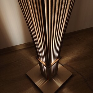 May include: A wooden floor lamp with a unique design. The lamp features a square base and a rectangular midsection, both made of light-colored wood. The top section flares out, composed of vertical wooden slats that allow light to filter through, creating a warm glow.