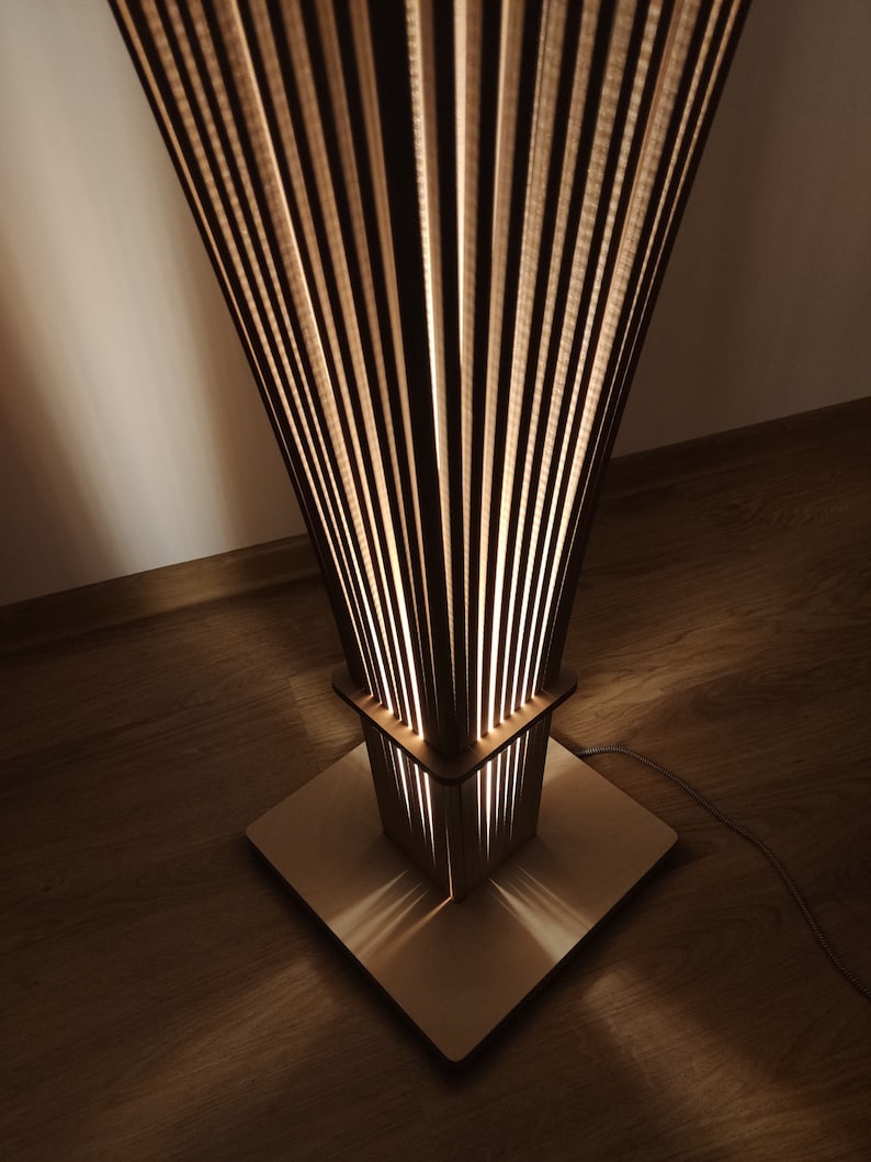 May include: A wooden floor lamp with a unique design. The lamp features a square base and a rectangular midsection, both made of light-colored wood. The top section flares out, composed of vertical wooden slats that allow light to filter through, creating a warm glow.