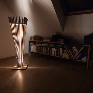 May include: A modern wooden floor lamp with a unique design. The lamp features vertical wooden slats that create a cone shape, with a square base and a cylindrical midsection. The lamp emits a warm, inviting glow, perfect for home decor.