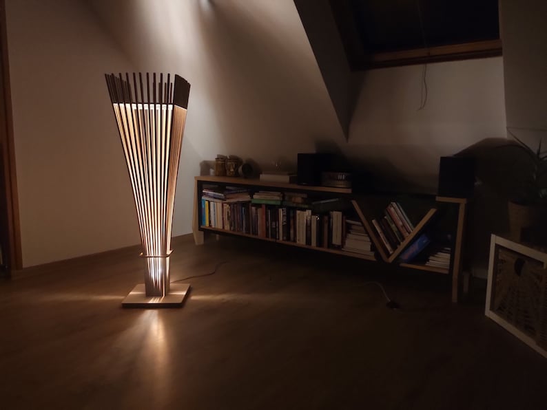 May include: A modern wooden floor lamp with a unique design. The lamp features vertical wooden slats that create a cone shape, with a square base and a cylindrical midsection. The lamp emits a warm, inviting glow, perfect for home decor.