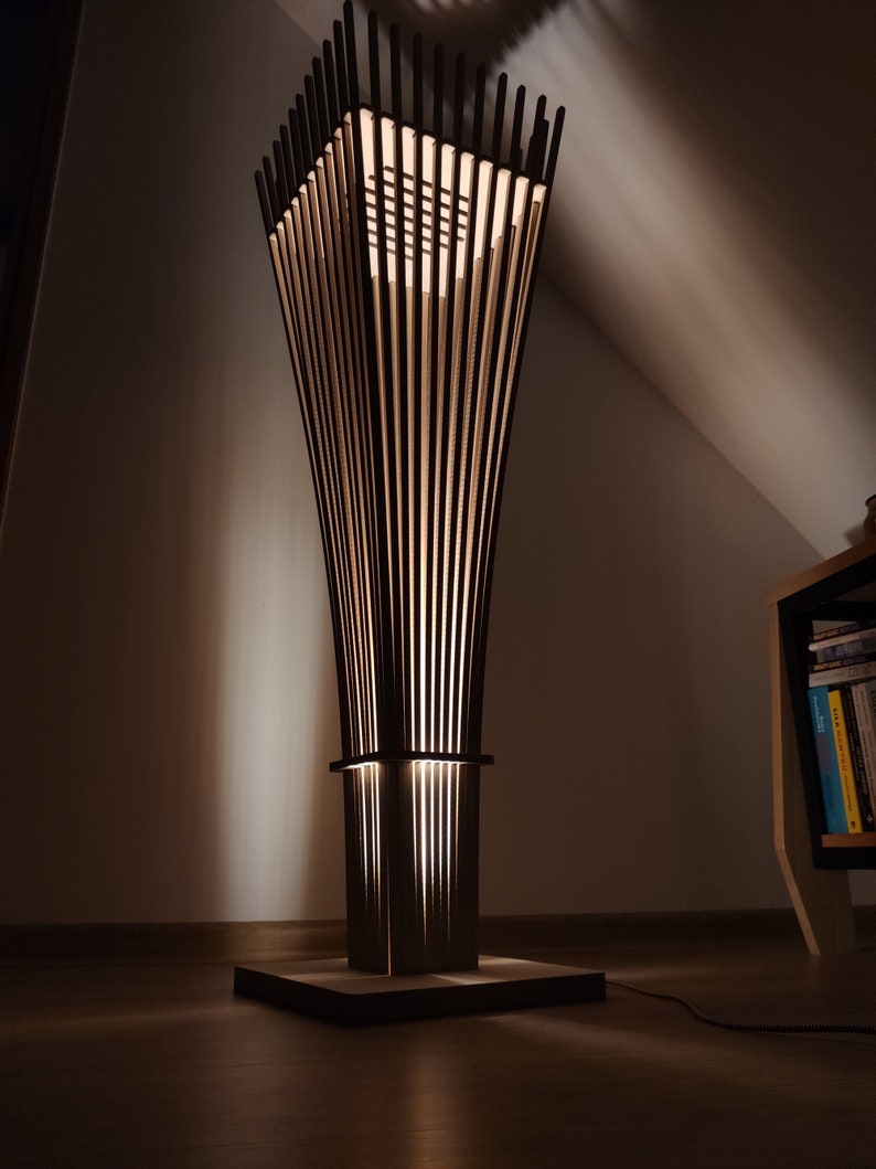 May include: A modern, wooden floor lamp with a unique design. The lamp features a square base, a rectangular midsection, and a flared top, all constructed with vertical wooden slats. The lamp emits a warm, inviting glow, creating a cozy ambiance.