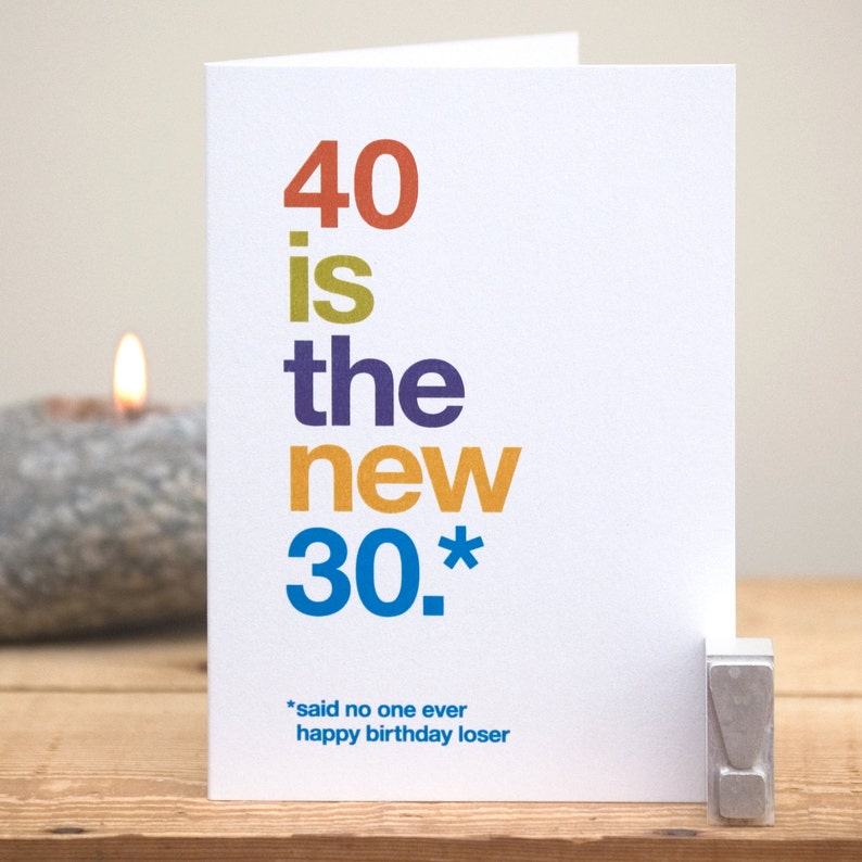 May include: A white birthday card with the text "40 is the new 30." in various colors. Below the text is "*said no one ever happy birthday loser." A lit candle and a gray stone candle holder are in the background.