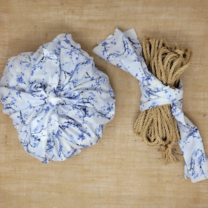 May include: Two items are displayed on a tan surface. One is a bundle wrapped in a white cloth with blue floral patterns. The other is a coil of brown rope tied with a matching cloth. The cloth has a blue floral pattern.