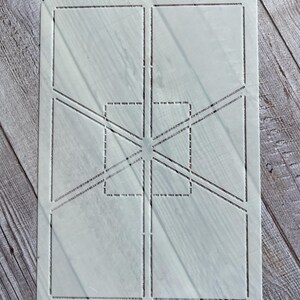 May include: A white plastic stencil with a geometric design. The stencil features rectangular and square shapes, along with diagonal lines and a central point. The design is cut out, allowing for paint or other mediums to be applied through the openings.