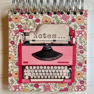 May include: A spiral-bound notepad featuring a pink typewriter design with the word "Notes..." printed on a paper. The notepad has a floral pattern background with pink, yellow, and green flowers.