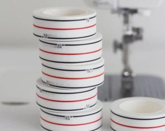 May include: White sewing tape rolls with black and red lines, marked with "¼" inch measurements. One stack of rolls is next to a single roll and a sewing machine in the background. Ideal for sewing and crafting projects.