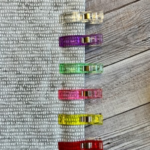 May include: Seven colorful plastic clips for sewing or quilting. The clips are arranged vertically on a gray fabric background with a white dot pattern. The clips are clear, purple, green, pink, yellow, and red.