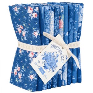 May include: A bundle of blue fabric squares with floral patterns and a white ribbon. The fabric features various shades of blue with pink and white floral designs. A tag reads "Something Blue" and "Tilda Edition".