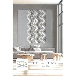 May include: A large, gray and white patterned wall hanging with a zigzag design hangs above a gray sofa. The text "ZEN CHIC" and "SHADOW PATH" are visible on a white square in the foreground. Two small wooden stools and a wooden coffee table are in front of the sofa.