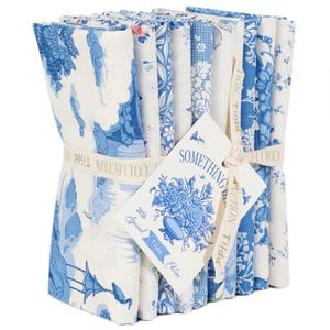 May include: A stack of blue and white patterned fabric squares tied with a cream ribbon. The fabric features floral and scenic designs. A tag with the text "SOMETHING DELICIOUS" and "Tilda Edition" is attached.