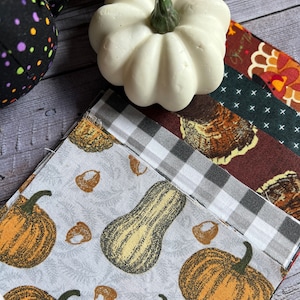 May include: A stack of fabric squares with various autumnal patterns, including pumpkins, gourds, and turkeys. The fabric squares are arranged in a staggered pattern, with the top square featuring a white pumpkin.