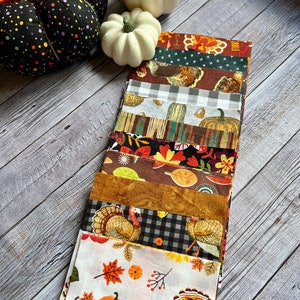 May include: A stack of 10 fabric squares with various Thanksgiving-themed patterns, including turkeys, pumpkins, leaves, and plaid. The squares are arranged in a vertical stack, with the top square being a red and brown fabric with a turkey design.