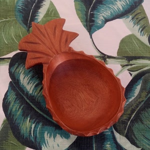 Könnte beinhalten: Eine hölzerne Schale in Ananasform mit einer rotbraunen Oberfläche. Die Schale ist mit einem detaillierten Ananas-Design geschnitzt und hat einen glatten, abgerundeten Innenraum.
