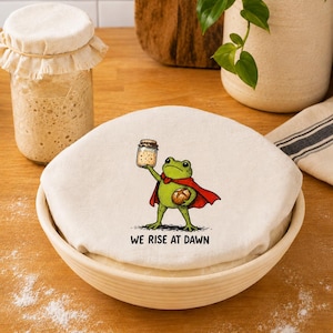 May include: A round, off-white fabric proofing bowl cover featuring a graphic illustration of a green frog wearing a red cape. The frog holds a glass jar and a loaf of bread. The text "WE RISE AT DAWN" is printed in black below the frog.