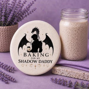 May include: A round, off-white fabric coaster featuring a black silhouette of a winged figure holding a jar, with the text "BAKING FOR MY SHADOW DADDY" printed in black below the figure, accented by small gold stars and decorative flourishes.