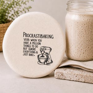 May include: A round, off-white fabric cover featuring black text that reads PROCRASTIBAKING VERB: WHEN YOU HAVE A MILLION THINGS TO DO BUT IGNORE EVERYTHING & JUST BAKE. Below the text is a black line drawing of a stand mixer with a mixing bowl.