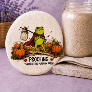 May include: A round, off-white fabric cover featuring a green frog wearing a red cape and carrying a jar on a stick. The frog walks through a pumpkin patch with orange pumpkins and autumn leaves. The text reads: PROOFING THROUGH THE PUMPKIN PATCH.