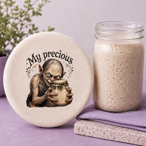 May include: A round, off-white fabric cover featuring a printed illustration of a pale, thin creature with large eyes holding a glass jar. The text "My precious" is printed in a dark, stylized font above the creature, surrounded by small leaf accents.