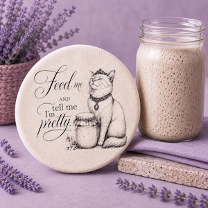 May include: A round, beige fabric-covered jar lid featuring a black ink illustration of a cat wearing a crown and necklace next to a jar. The text reads, "Feed me AND tell me I'm pretty." The lid is positioned beside a glass jar filled with beige liquid.