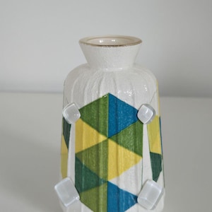 Pode incluir: Um vaso de cerâmica branco com uma superfície texturizada e um design geométrico em amarelo, verde e azul. O vaso tem um pescoço estreito e é adornado com embelezamentos quadrados transparentes.