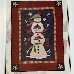 May include: A framed fabric art piece titled "Stack of Snowmen." The artwork features a stack of three snowmen with hats, scarves, and bird accents against a dark blue background. The frame is a dark red with snowflake and star accents.