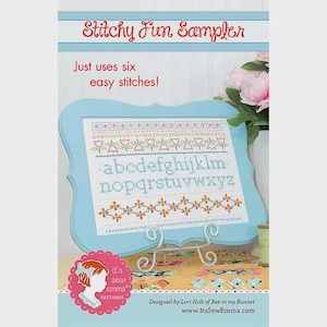 May include: A light blue framed cross-stitch sampler with the text "Stitchy Fun Sampler" and "Just uses six easy stitches!" The sampler features floral and alphabet patterns in various colors. A spool of thread and pink flowers are also visible.
