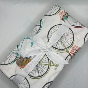 May include: A rectangular package wrapped in white fabric with a bicycle wheel pattern in black, teal, and red. The package is tied with a white satin ribbon and bow. The background is white.
