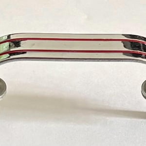 May include: A chrome-finished cabinet pull handle with three red accent lines. The curved handle has two mounting posts. This hardware is suitable for drawers, cabinets, or other furniture.