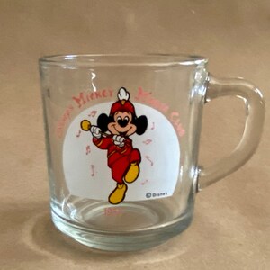 May include: A clear glass mug with a handle. The mug has a white circle with a cartoon image of Mickey Mouse playing a trumpet. The text "Disney's Mickey Mouse Club" is printed around the circle in pink.