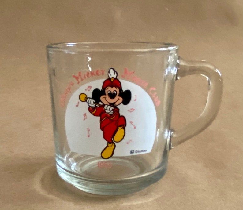 May include: A clear glass mug with a handle. The mug has a white circle with a cartoon image of Mickey Mouse playing a trumpet. The text "Disney's Mickey Mouse Club" is printed around the circle in pink.