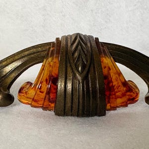 May include: A drawer pull featuring a central amber-colored translucent resin element with a leaf-like pattern, flanked by two curved, dark bronze-toned metal arms that extend outward with decorative, rounded, fan-shaped ends.