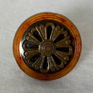 May include: A circular button featuring a translucent amber-colored outer rim and a dark brown center. The center is inlaid with a metallic bronze-toned floral motif consisting of radiating petals and a small circular detail at the very middle.