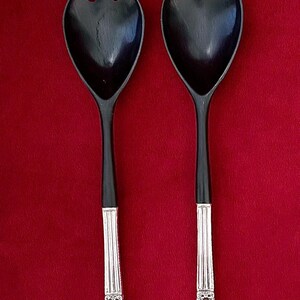 May include: A pair of black salad servers with silver-colored handles. One server has a fork-like end, the other a spoon-like end. The handles have a vertical ribbed design and decorative floral elements.