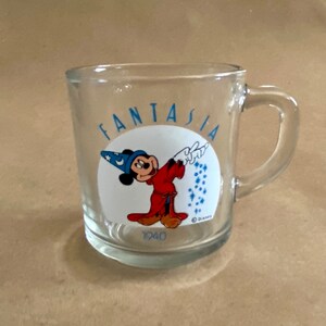 May include: A clear glass mug with a handle. The mug has a white circle with a cartoon image of Mickey Mouse dressed as the Sorcerer's Apprentice from the movie Fantasia. The text "Fantasia" is written around the circle in blue letters. The text "1940" is printed below the circle and the text "&copy; Disney" is printed below that.