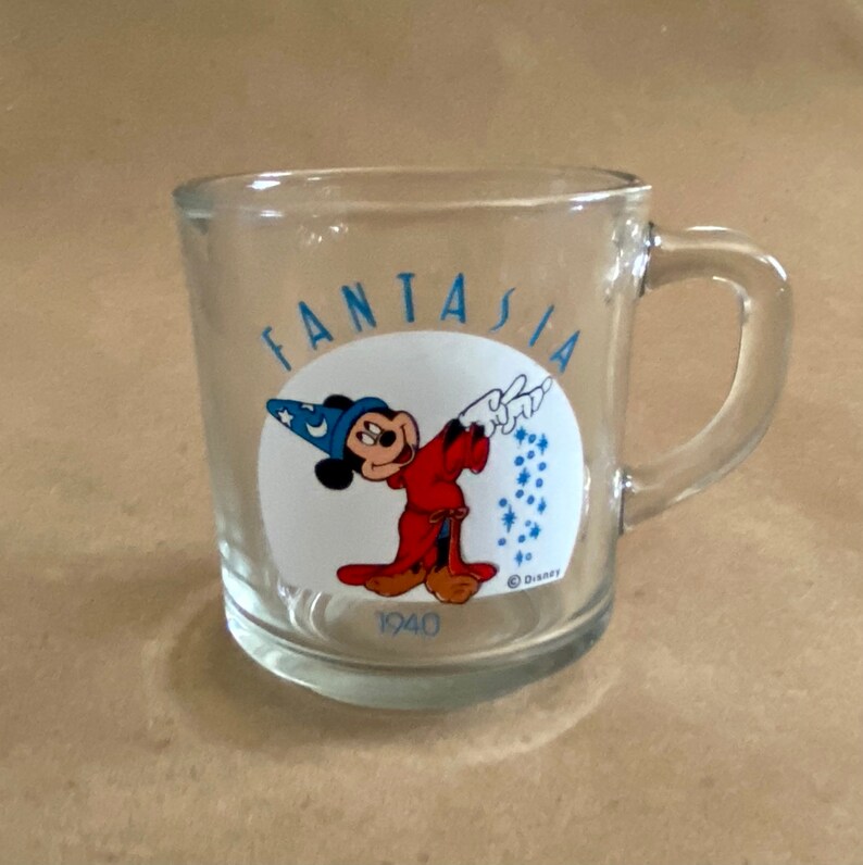 May include: A clear glass mug with a handle. The mug has a white circle with a cartoon image of Mickey Mouse dressed as the Sorcerer's Apprentice from the movie Fantasia. The text "Fantasia" is written around the circle in blue letters. The text "1940" is printed below the circle and the text "&copy; Disney" is printed below that.