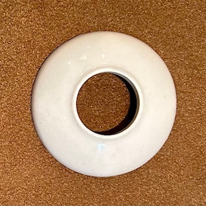May include: A white, circular ceramic object with a hole in the center. The object has a smooth, glossy surface and appears to be a decorative or functional item. The item is set against a brown background.