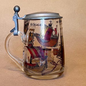 May include: A glass beer stein with a pewter lid and a handle. The stein is decorated with a colorful illustration of ships, including a Viking longship and a cog, with the word "KOGGE" visible on the side.