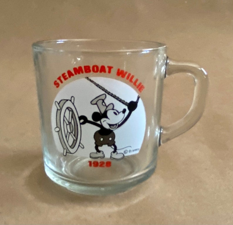 May include: A clear glass mug with a handle. The mug has a black and white image of Mickey Mouse as Steamboat Willie with the text "Steamboat Willie 1928" and a Disney copyright symbol.
