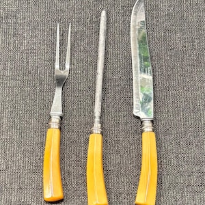 May include: A set of three vintage kitchen utensils with yellow plastic handles. The set includes a fork, a knife, and a sharpening steel.