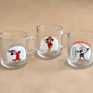 May include: Three clear glass mugs with handles. Each mug features a different image of Mickey Mouse from classic Disney cartoons. The first mug shows Mickey Mouse as a sorcerer, the second mug shows Mickey Mouse as a bandleader, and the third mug shows Mickey Mouse as Steamboat Willie.