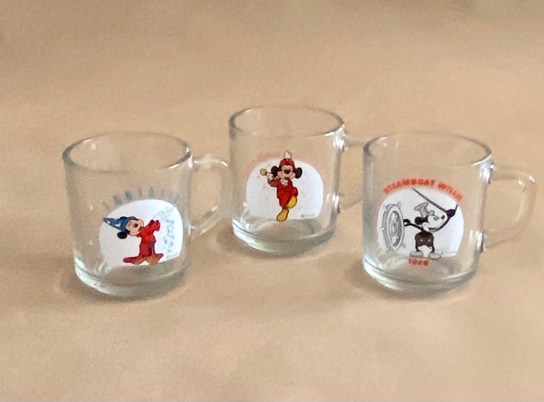 May include: Three clear glass mugs with handles. Each mug features a different image of Mickey Mouse from classic Disney cartoons. The first mug shows Mickey Mouse as a sorcerer, the second mug shows Mickey Mouse as a bandleader, and the third mug shows Mickey Mouse as Steamboat Willie.