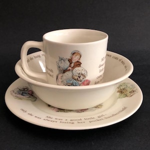 May include: A white ceramic children's dish set featuring illustrations and text. The set includes a mug, a bowl, and a plate. The illustrations depict a girl and a dog. Text includes phrases like "She was a good little girl" and "All down coats of mice".