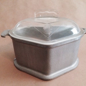 May include: A square, gray metal casserole dish with a clear glass lid. The lid has a decorative handle and etched design. The dish has two side handles and a textured exterior. A vintage kitchenware item.