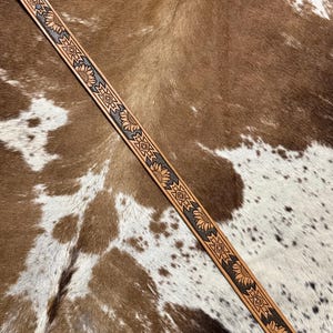 May include: A light brown leather belt with a sunflower and geometric pattern. The belt is approximately 1.5 inches wide and features a detailed embossed design. The belt is displayed on a brown and white cowhide background.